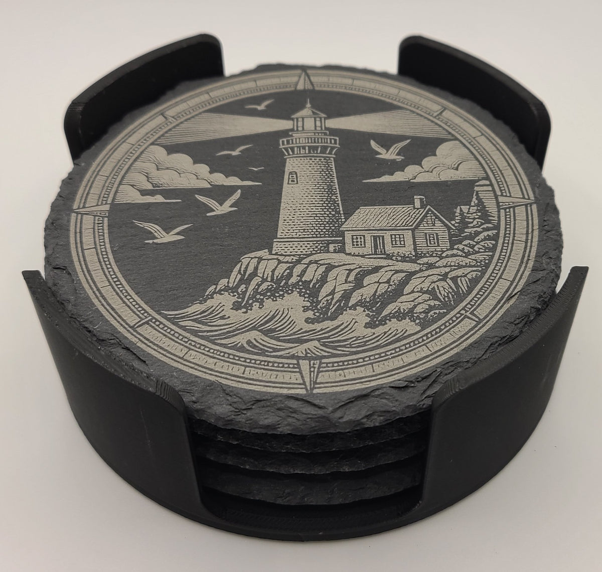 A set of round slate coasters with a black plastic ring holder, featuring an engraved nautical scene of a lighthouse, waves, clouds, and flying seagulls.