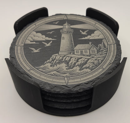A set of round slate coasters with a black plastic ring holder, featuring an engraved nautical scene of a lighthouse, waves, clouds, and flying seagulls.